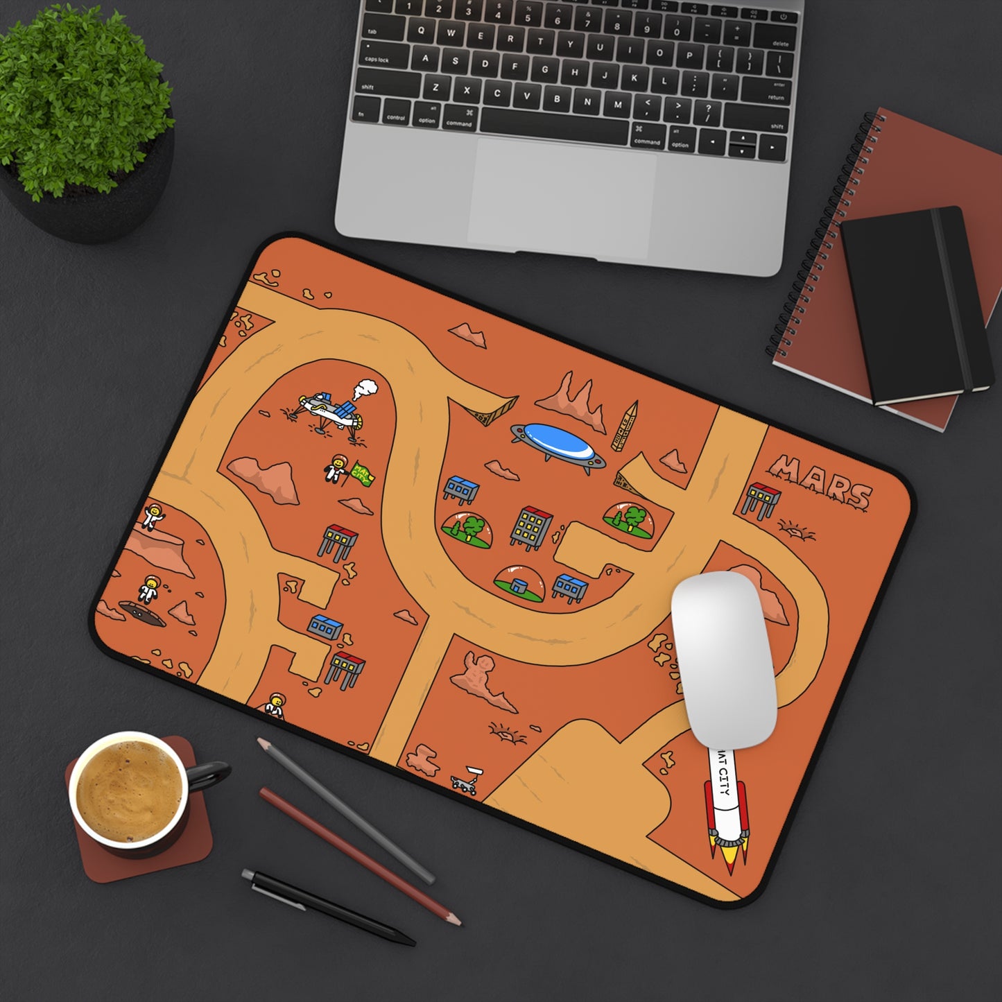 The Matcity Mars Mouse Mat Medium on a dark desk setup with a laptop, notebook, and a cup of coffee. The design adds color and creativity to the workspace, highlighting the detailed Martian city.