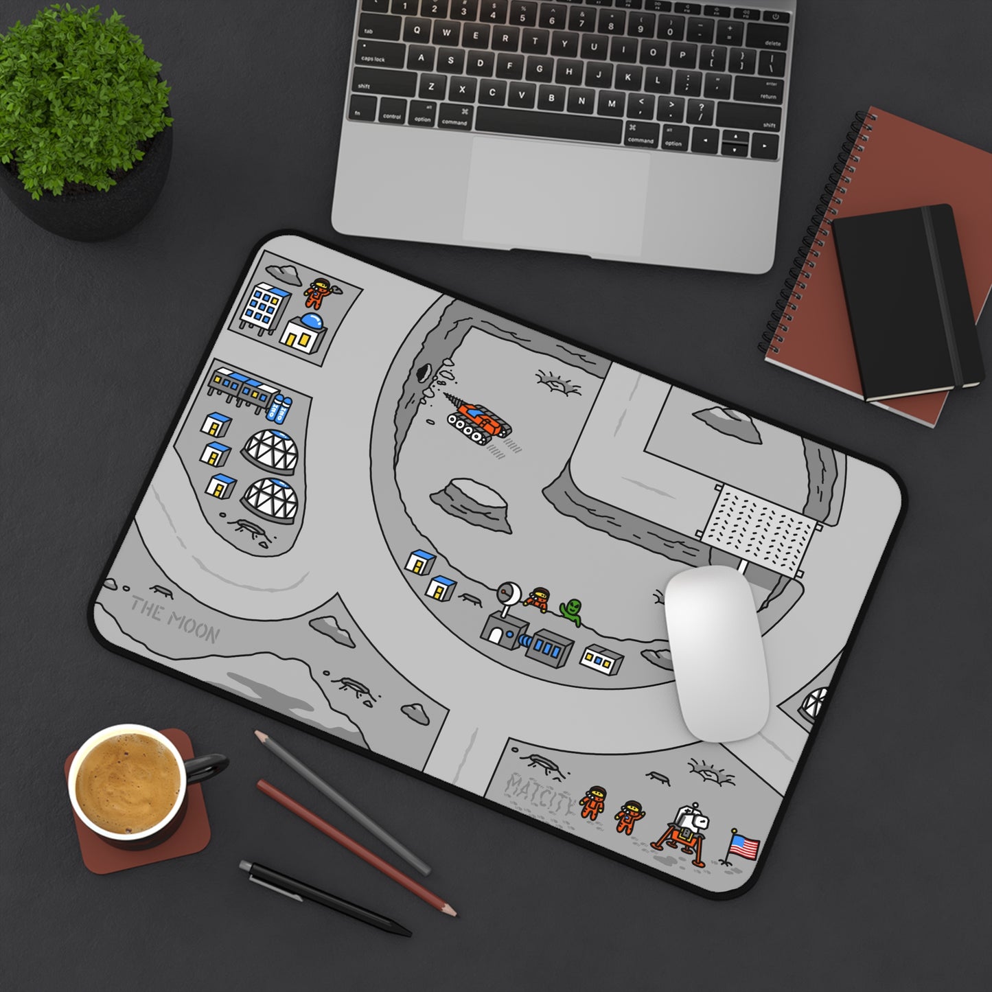 Matcity Moon Mouse Mat Medium on a dark desk with a laptop, plant, notebook, and coffee. The mat’s design shows lunar habitats, astronauts, and vehicles, creating a playful space-themed workspace.