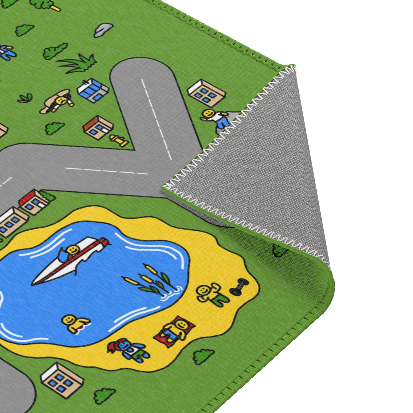 A close-up view of the Matcity play mat, showing the corner where the mat is folded to reveal its underside with detailed stitching. The vibrant design on the front features a section of a road near a pond with a boat and small characters relaxing on a yellow beach. The colors are bright and playful, with green grass, blue water, and various small buildings scattered around the mat.